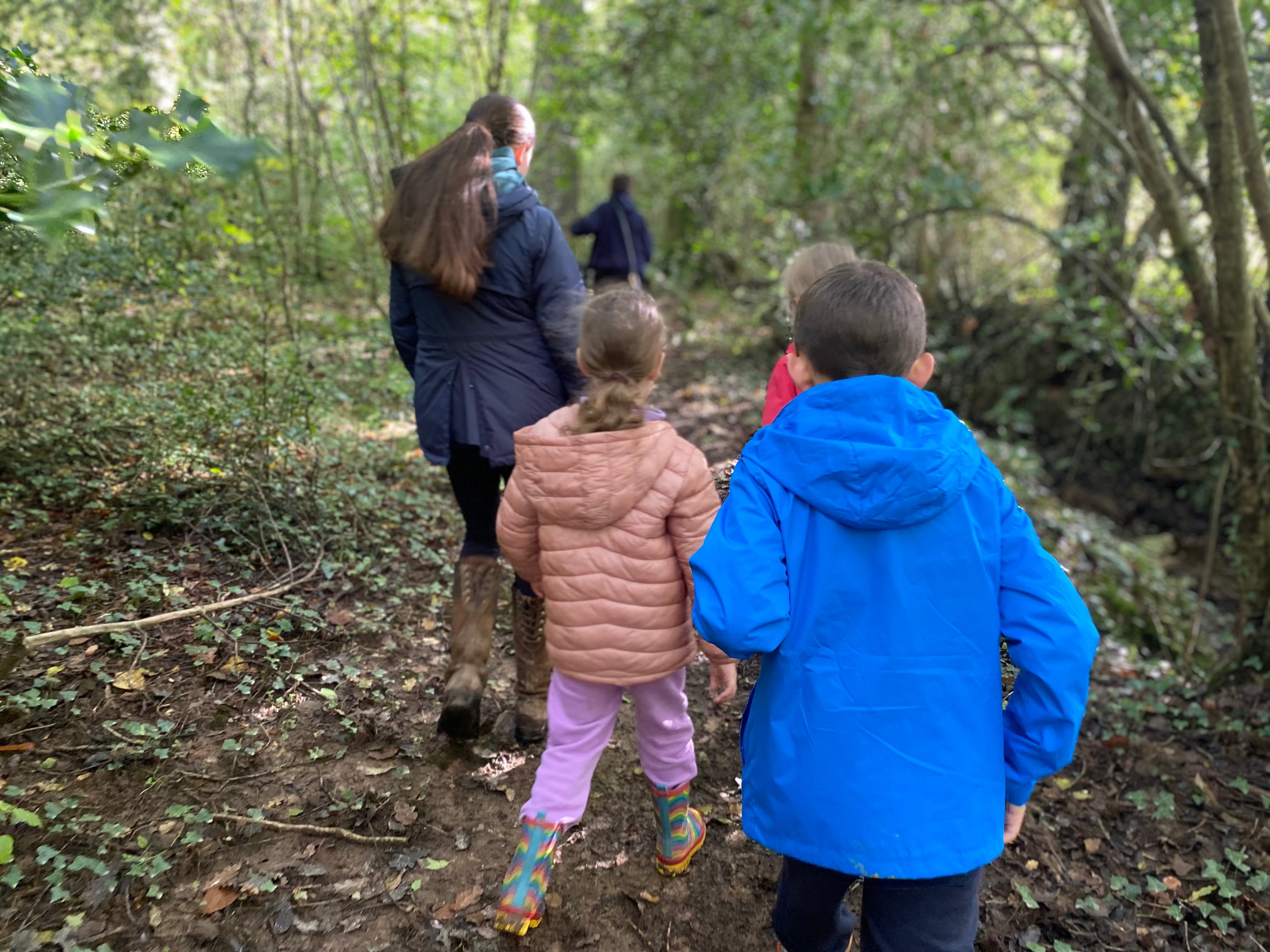 A group of yound people are walking through the woods, there are four young people and a woman. A girl of about seven is wearing a coral coloured puffa jacket and pink jeans, a boy of about 10 in a blue jacket and black jeans. We can see another couple of children in the distance.