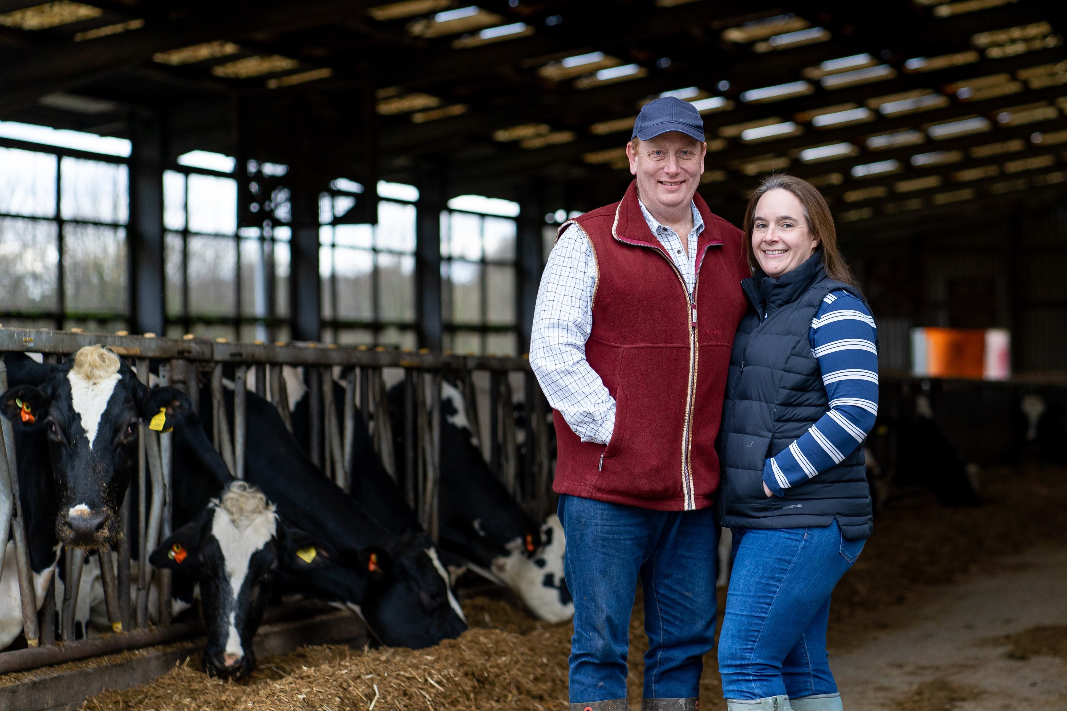 Two farmers stand in a milking shed. They are Nick and Lucy Tyler of Kingsplay Farm. Black and white cows nibble on hay behind them in the milking stalls.