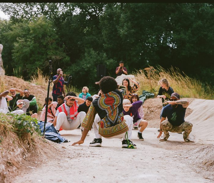 A group of people are dancing in Luton henge, one has his back to use and the others are facing us. They've all got down low into a crouch with one hand behind them on the floor and the other across their forehead. The dancing might be Bollywood dancing. They are all enjoying themselves and we can see one of the Luton Henge standing stones in the background towards the top left of the picture. There are trees in the background.