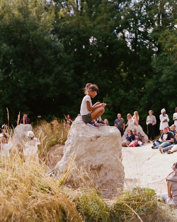 A little girl of about seven is sitting on top of one of the wider Luton Henge standing stones - the stone is light grey. The girl is wearing dark shorts and a white t-shirt. Other people are sitting on the floor in the distance. The floor is made of compacted white chalk, part of the henge. There are trees in the background.