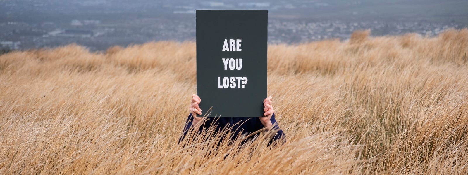 A man is crouching in long brown grass holding a large black sign obscuring his face, it says, in white, Are You Lost?