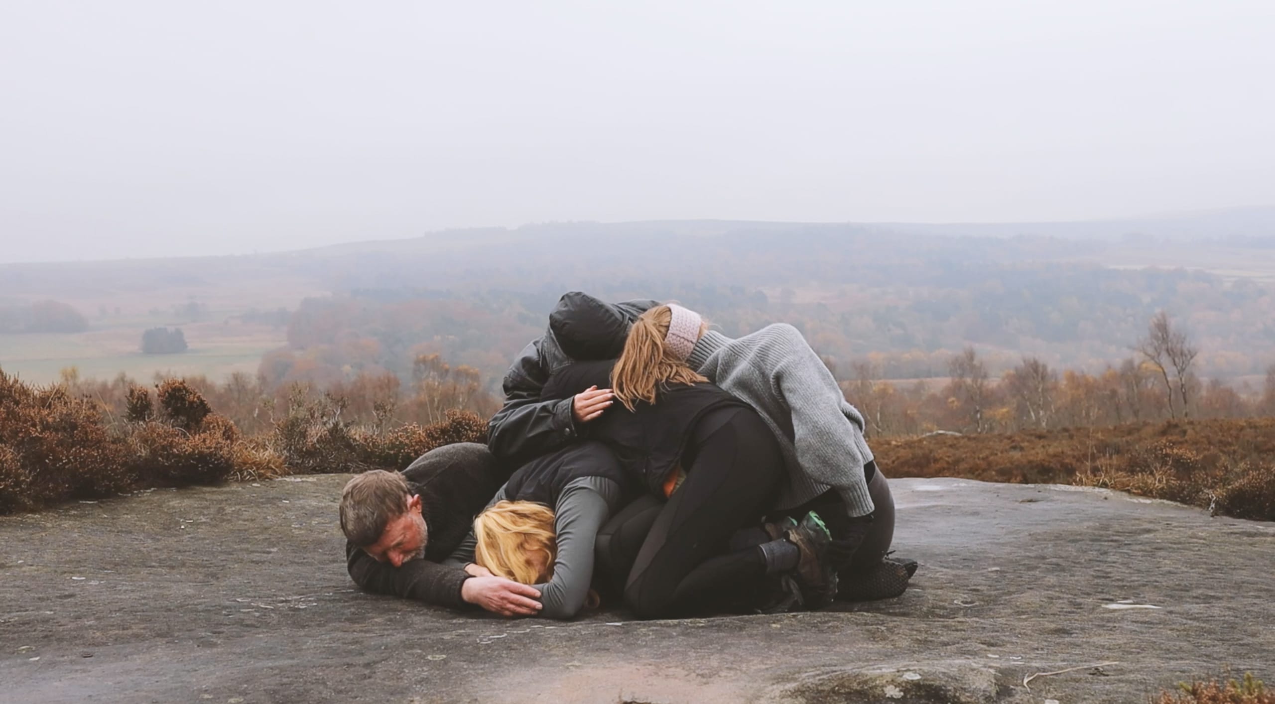 Dancers folded up together on a large flat rock with a misty landscape in the background.