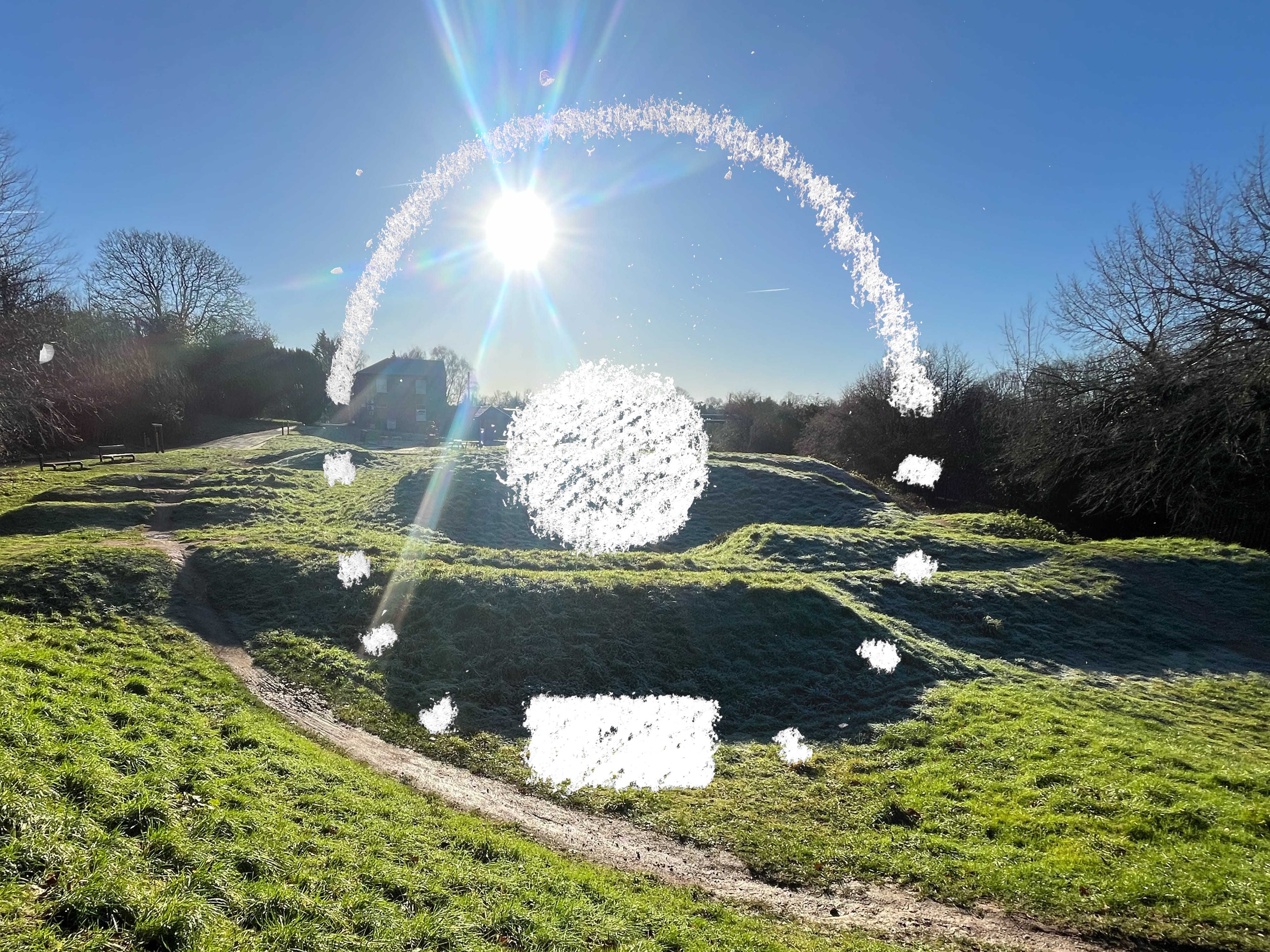 A view of a lumpy landscape! There are grass covered earthworks bordered by trees. The sun is shining in a cloudless sky. Superimposed on the image is a series of chalk symbols, a large dot in the middle with a semi circular line above it, following the same arc. Making up the rest of the circle is a series of small dots and a filled in rectangle below the central dot.