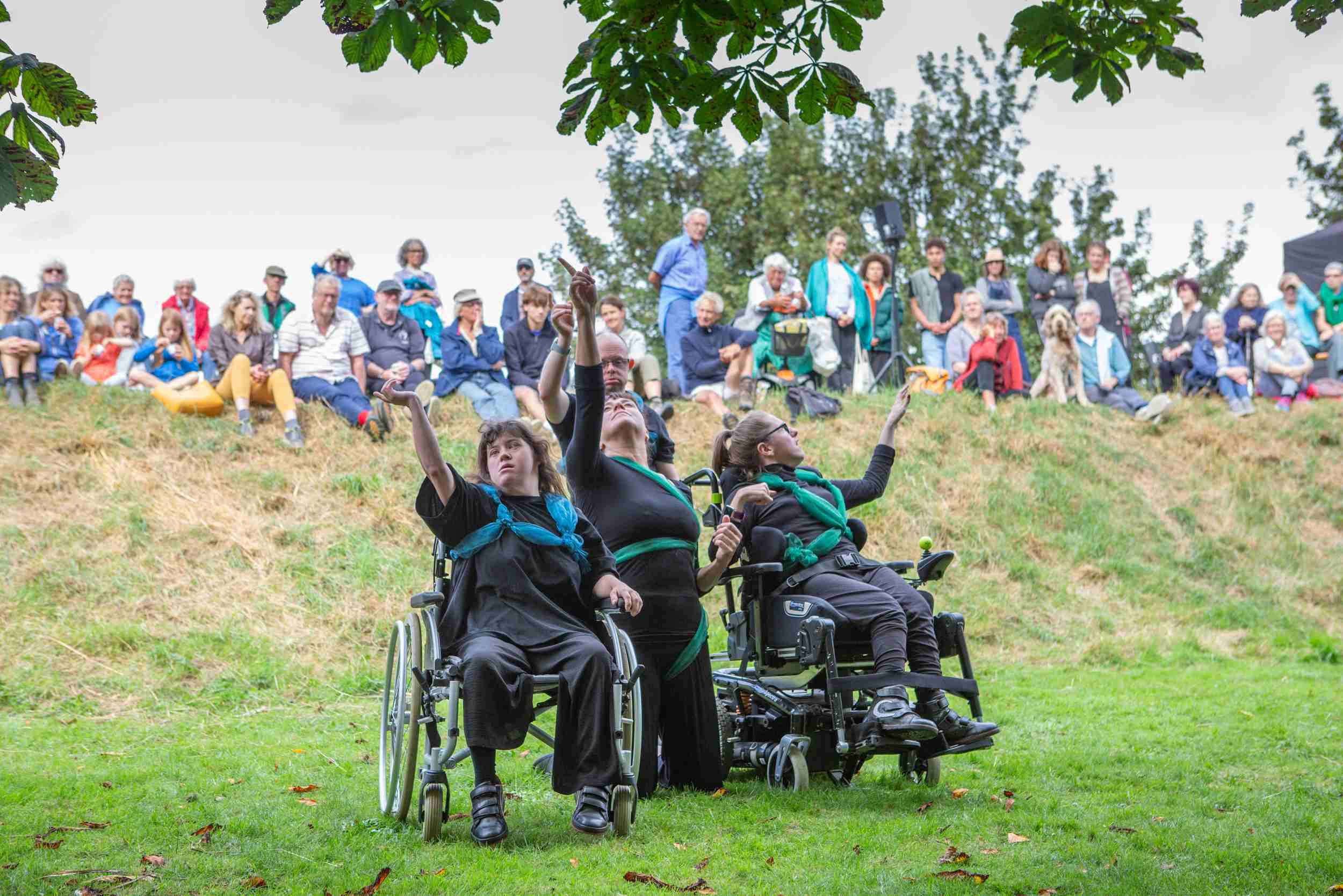 Two people in wheelchairs and one person kneeling hold their hands in the air, dancing for a crowd watching them from on top of a knoll.