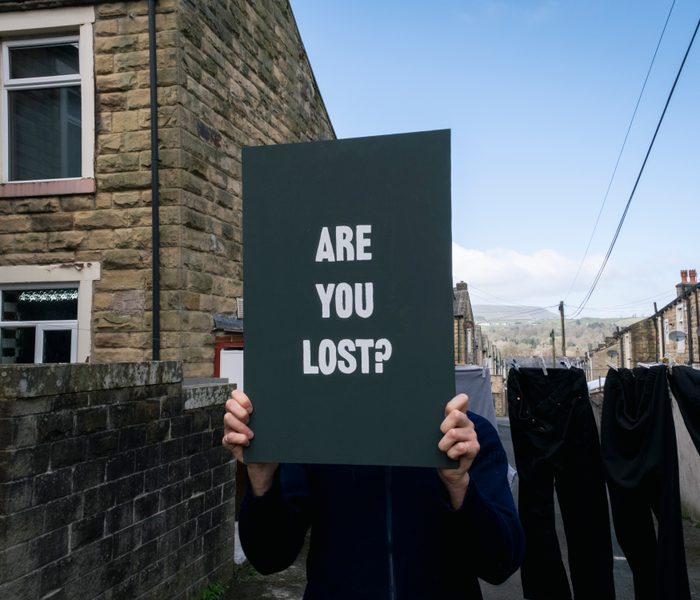 A man stands in a cobbled back street in Lancashire. There is a line of washing behind him. He is holding a sign in front of his face saying Are you Lost? in white capital letters.