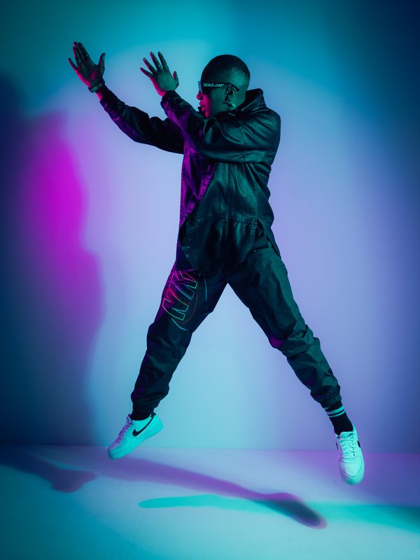 A woman jumps in the air, almost levitating. She looks to the left, towards her outstretched hands. She's wearing shades and a black leather jacket. The lighting is purple and turquoise.