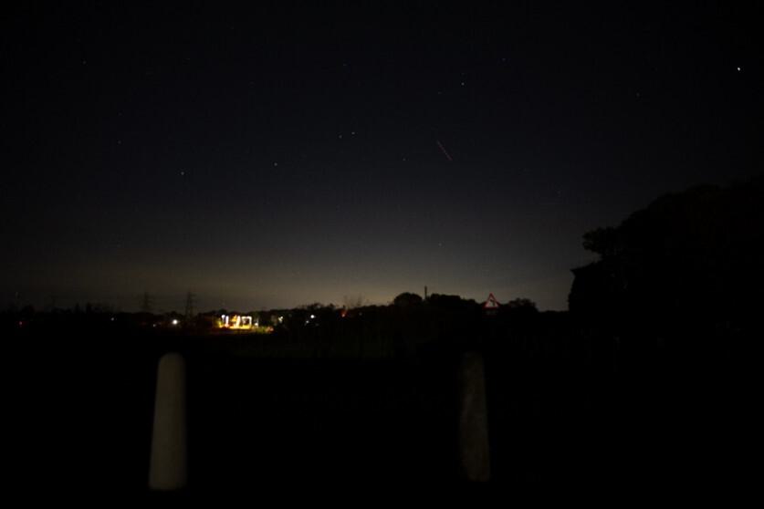 A night time scene, the sky is dark and we can see the silhouette of a tree on the right hand side. In the distance, on the left, a lit up structure dominates the image. This is Manningtree station