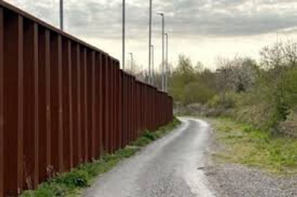 A thick metal fence snakes down the left hand side of the photo, it is about 10 feet tall, behind it are very tall football stadium style lights
