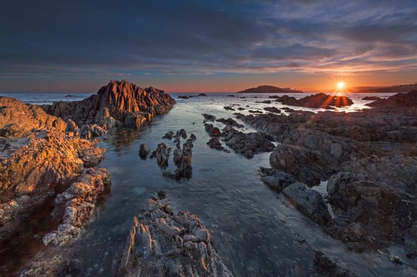 A very low sun - touching the horizon, sets over costal rocks.