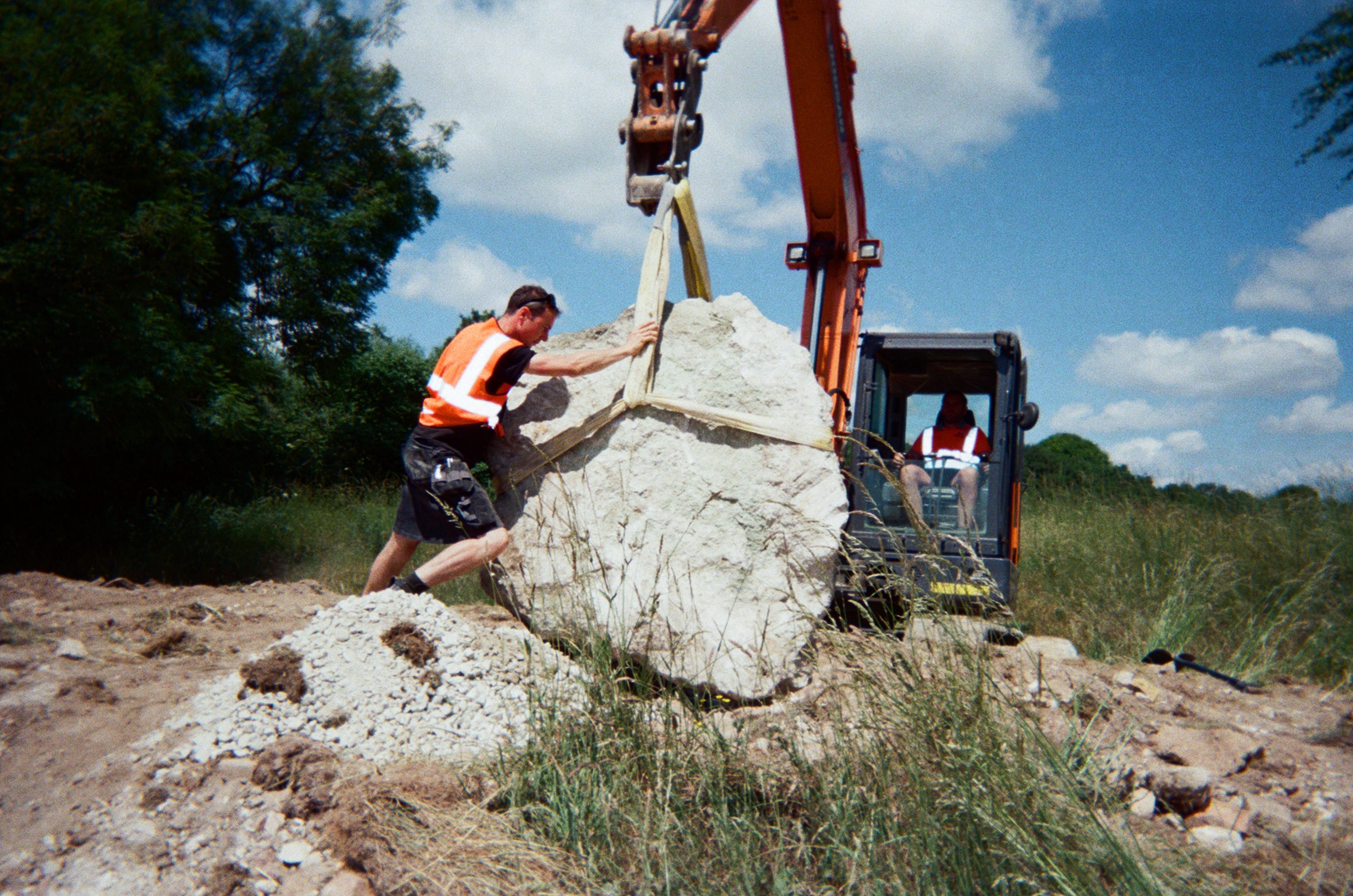 A man in an orange hi viz is helping guide a large rock into place. It is being lifted by an orange digger using a strap around the rock.