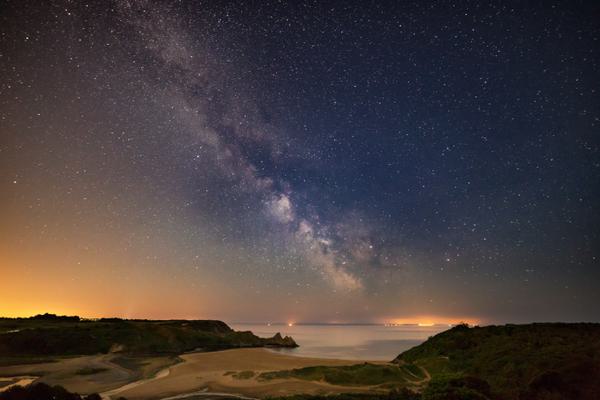 The milky way runs from the top left of the photo to the centre. The sky is orange at the horizon. Cliffs are silhouetted and the sea is in the distance.