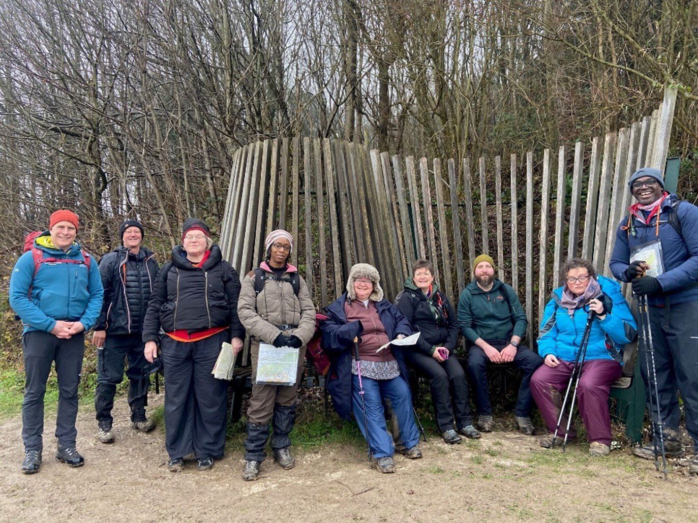 Nine people smile at the camera, they are wearing warm outdoor clothes and woolly hats. Some of them are standing, others sitting on a bench with a fence and trees behind.