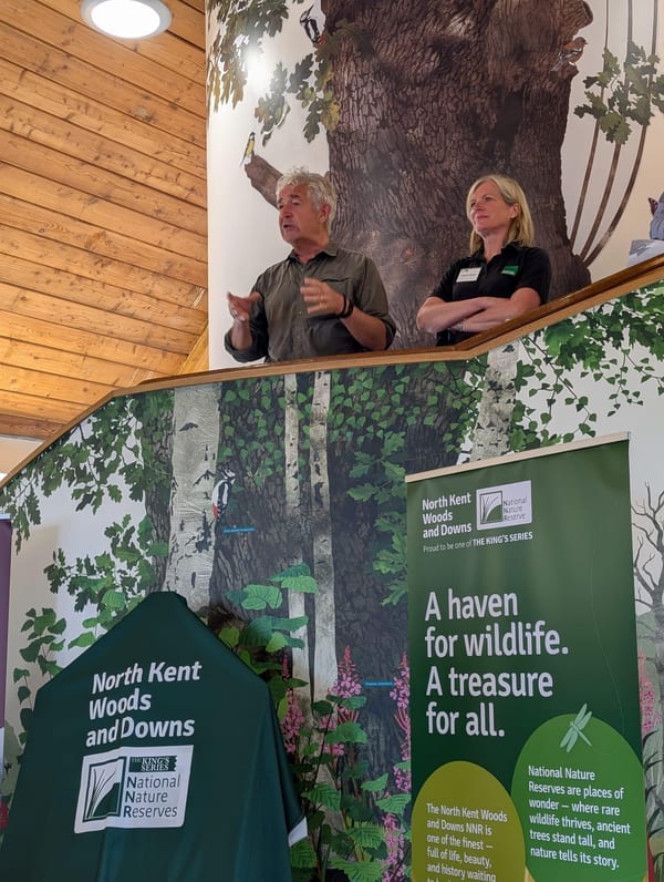 Tony Juniper and Marian Spain are speaking on an elevated landing. Tony has grey hair and wears a shirt, Marian has blonde shoulder length hair and wears a Natural England polo top. The landing has National Nature Reserve branded flags in front of it and graphics of nature.