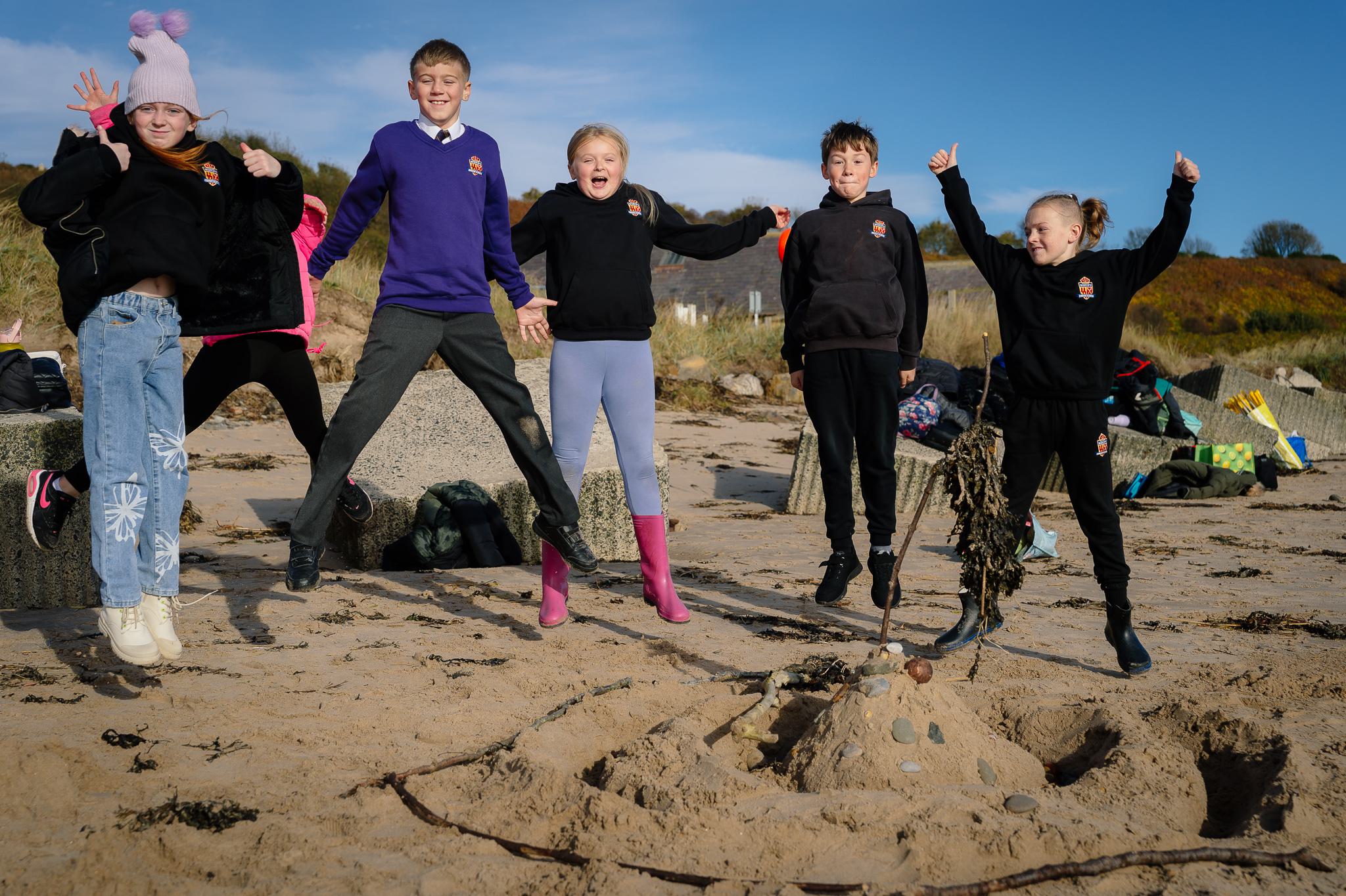Six children leap into the air! They are about ten years old. They're on the beach behind a sandcastle they have built. They look very happy