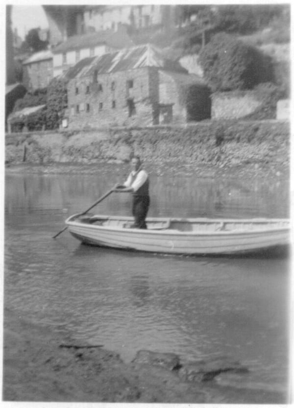 A man is propelling a small passenger ferry with a pole. It's a black and white image from the 1940s and we can see an old warehouse in the background.