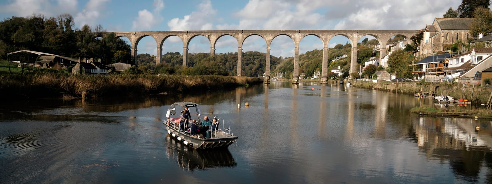 A long shot of a small passenger ferry with passengers aboard and the iconic Calstock Viaduct in the background. It's a tall structure made of stone with very high arches.