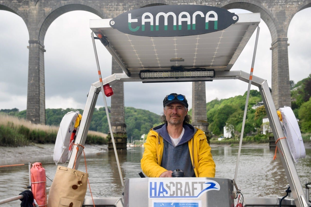 A man drives a boat, he's standing under an awning with solar panels on top and the word Tamara across the front. In the background we can see the very tall Calstock Viaduct. He is wearing a yellow jacket and a beany hat. He has shoulder length hair and a moustache.