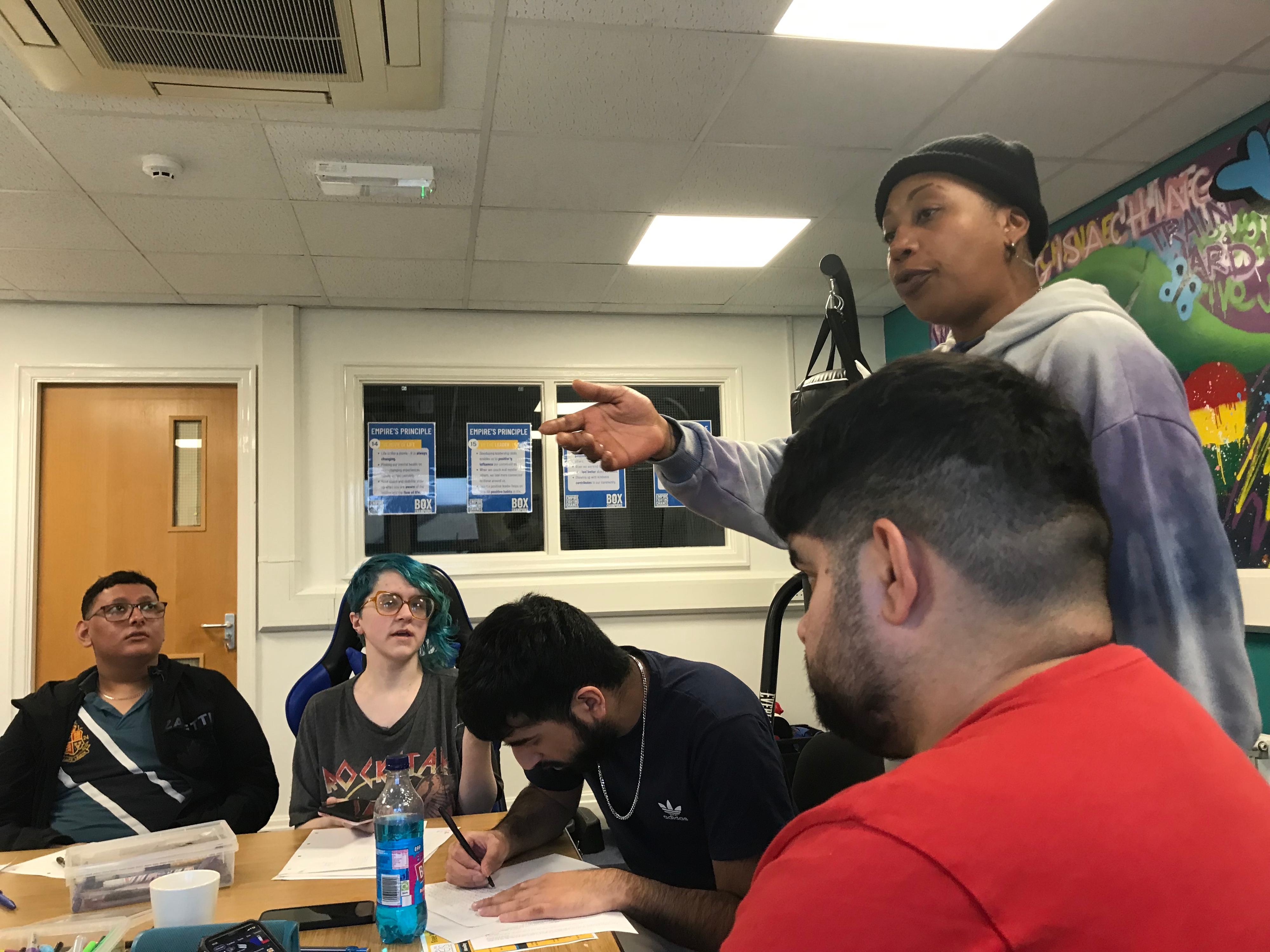OneDa is facilitating a workshop with young people. Some of them are writing their ideas down. Some of them are from South Asian backgrounds, others are white. One has blue and green hair. OneDa has her hand pointing forward like she is sharing an idea. She is wearing a black beanie and a grey zip up hoodie.