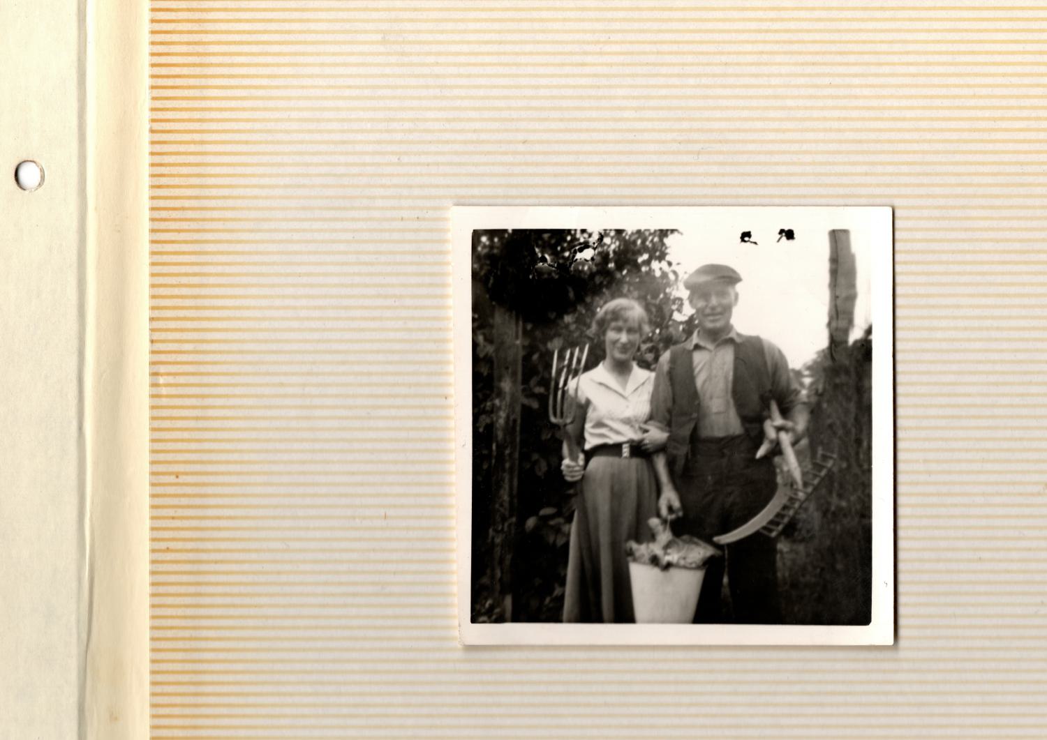 Sam and Beryl A small square photograph with a white trim, stuck to the page of a photo album. The page is covered in horizontal orange lines, faded and weathered with age. On the left hand side is a cream strip with a hole punch, designed to fit into an album. The photograph itself is black and white, and shows a young woman holding the arm of an older man. Behind them sits foliage, perhaps part of a farm. The young woman, Beryl, holds a pitchfork in one hand, and the arm of her father with the other. She wears a long circle skirt, belted with a large clasp, and a white shirt. Her hair is short and slightly curled. Sam wears a shirt and an open waistcoat. He holds a bucket of what may be cabbages in one hand, and a scythe and rake in the other. His trousers are high waisted, his shirt is tucked in, and atop his head sits a flat cap. Both are smiling.