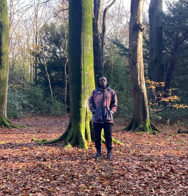 Still Shadey is standing in front of a tree looking directly at the camera. He has locks and is wearing a jacket and a pair of black jeans. He looks thoughtful. The tree is tall and straight and has moss on its trunk. The floor is covered in autumn leaves.