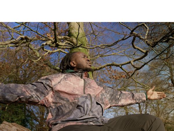 Still Shadey sits on a log. He has locks and is facing the right of the photo with eyes closed and arms held out wide. He looks thoughtful. Behind him are trees and a stormy blue sky.