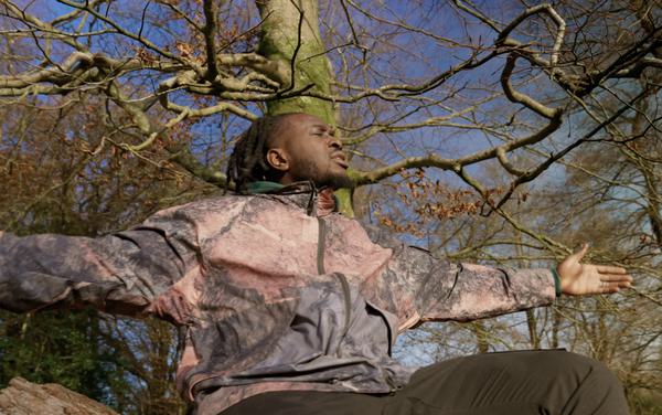 Still Shadey sits on a log. He has locks and is facing the right of the photo with eyes closed and arms held out wide. He looks thoughtful. Behind him are trees and a stormy blue sky.